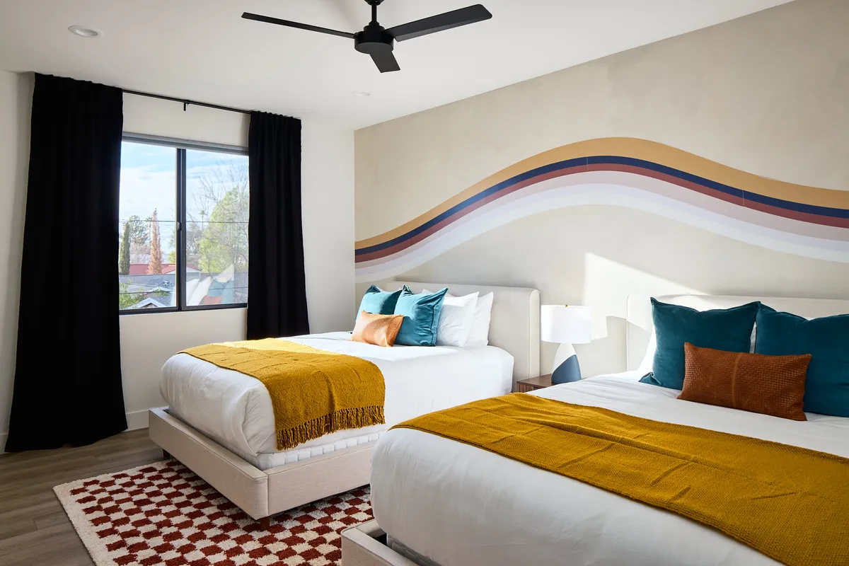 A cheerful upstairs bedroom featuring two beds with colorful throws and pillows, a bold abstract wall mural, and a large window letting in abundant natural light. The room's layout and playful decor evoke a sense of vibrancy and comfort, perfect for families or friends.