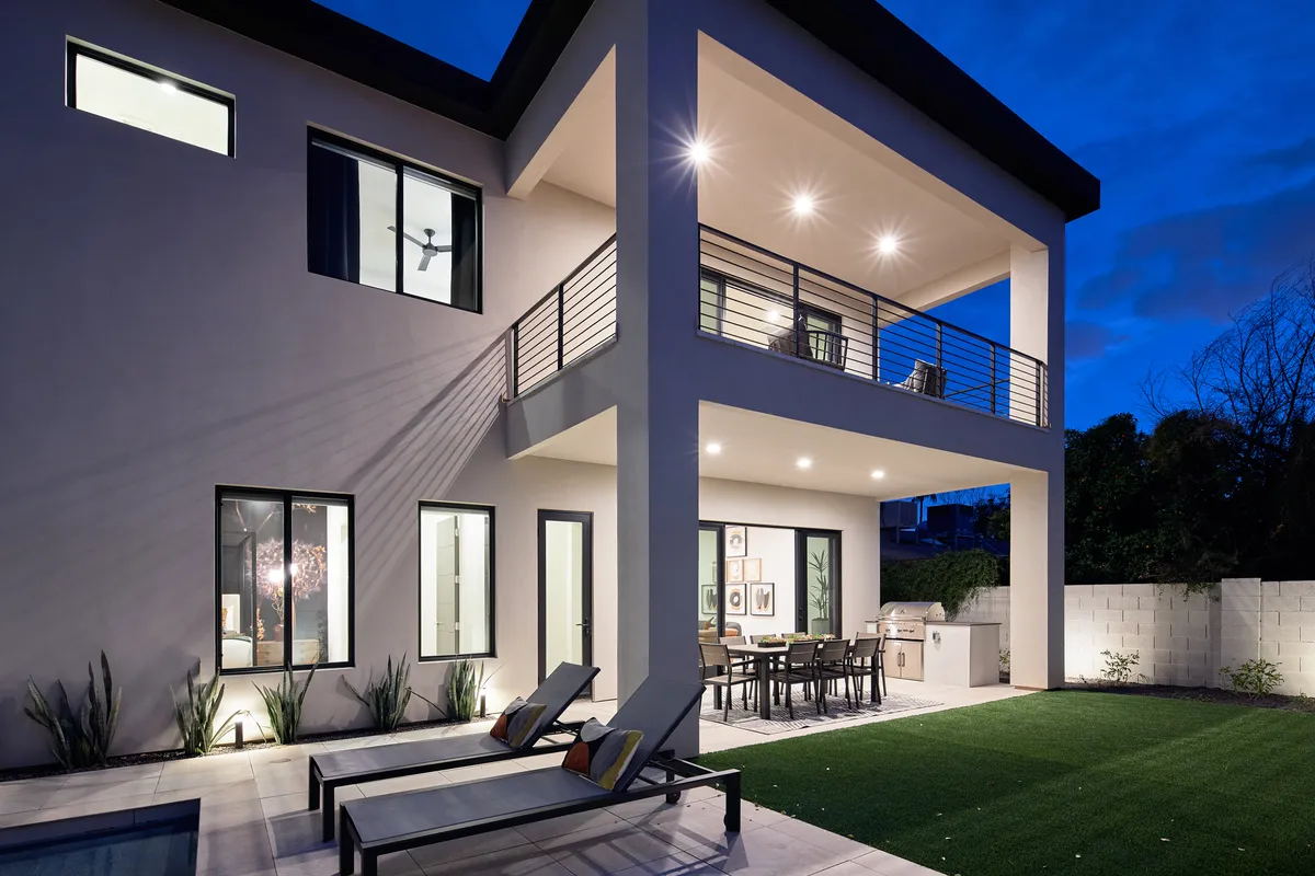 A wide exterior shot of a modern property featuring a sleek outdoor dining area, poolside lounge chairs, landscaped gardens, and an upper balcony. The setting is illuminated for evening gatherings, capturing the ambiance of resort-style relaxation and social entertainment.