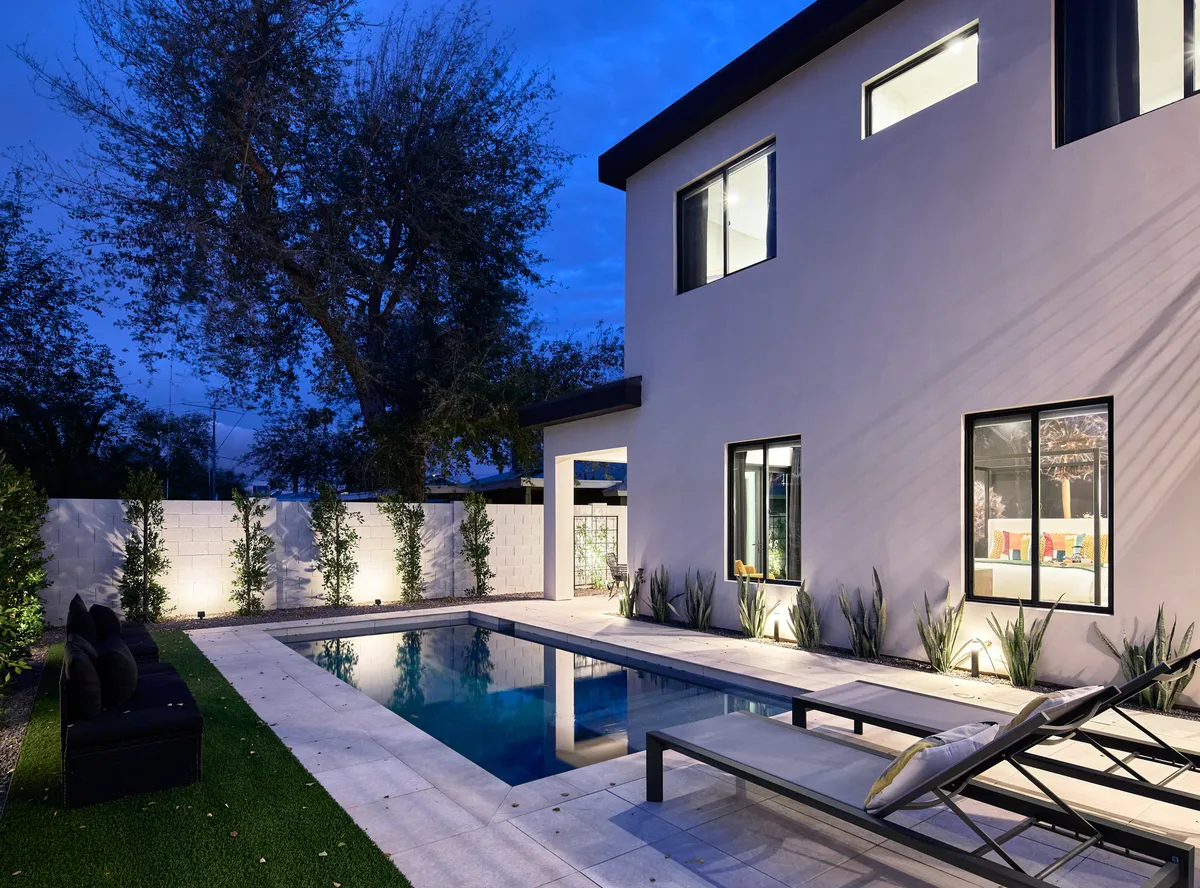 A serene evening view of a modern backyard with a swimming pool, surrounded by plush lounge chairs and lush landscaping. The tranquil atmosphere is elevated by subtle lighting and the sleek design of the home, ideal for sunset relaxation and intimate gatherings.