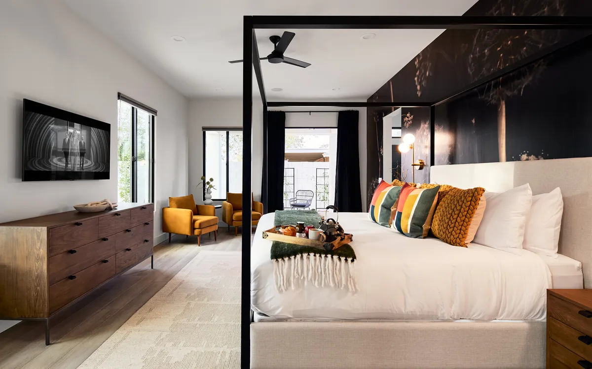 A spacious primary bedroom with a four-poster bed adorned with colorful pillows, a cozy seating area featuring yellow chairs, a wooden dresser, and large windows that open directly to an outdoor space, allowing natural light to flood the room. The inviting arrangement hints at both relaxation and easy access to the backyard for morning coffee.