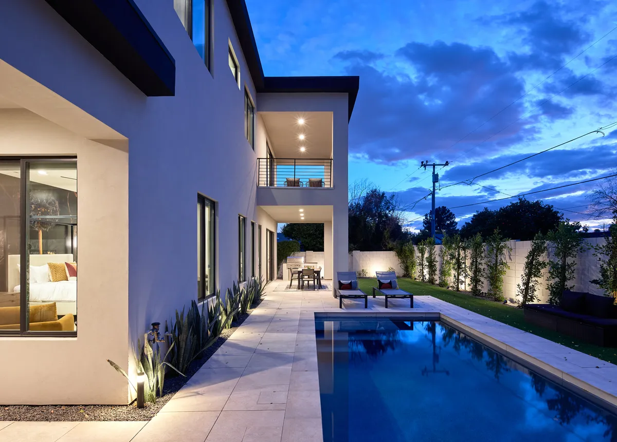 Showcases an inviting backyard oasis with a swimming pool, outdoor seating, lush landscaping, and large windows. The scene radiates relaxation, with hints of evening ambiance ideal for returning home after a day of exploring.