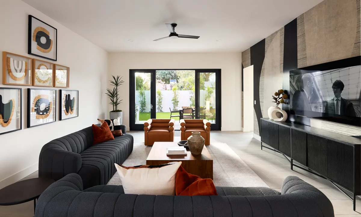 A bright, open-plan living room featuring a comfortable sofa, modern artwork, a coffee table, and a large television, with sunlight streaming through windows and a view to the outdoors. The decor blends contemporary style with boho-chic touches, creating an inviting space for relaxing or socializing.