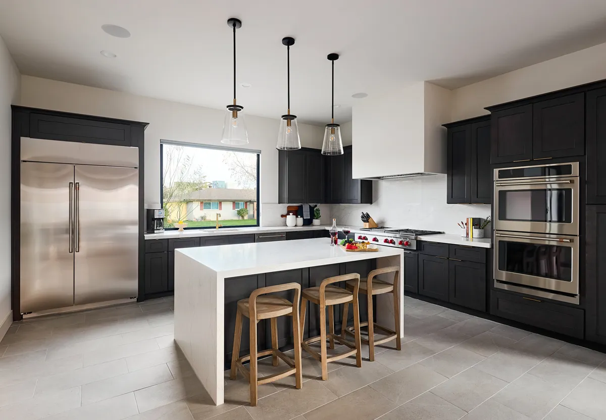 This image showcases a modern, open kitchen featuring a spacious island countertop, stainless steel appliances, stylish pendant lights, and a large window that provides plenty of natural light. The setting immediately communicates a space designed for both culinary creativity and social interaction.